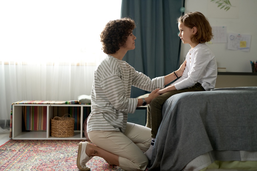 A woman kneeling beside a bed and holding a child's arms while they face each other in a bright bedroom.