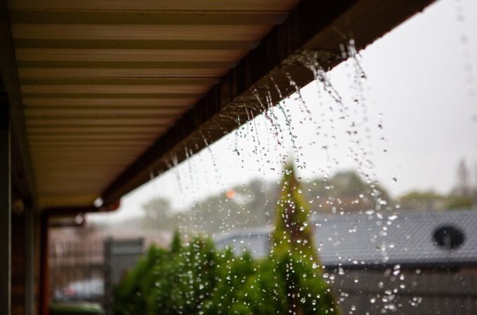 water spilling from a roof gutter in heavy rain, with droplets falling beside a covered house exterior.