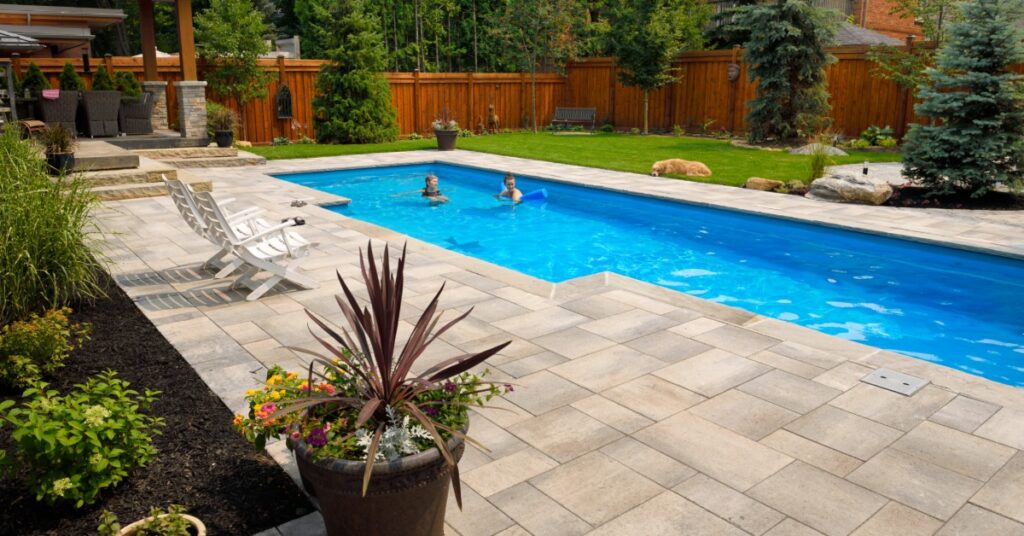 Two people swimming in a rectangular backyard pool surrounded by a stone patio, lounge chairs, and greenery.