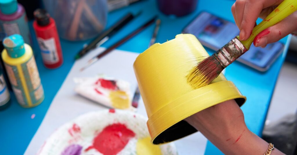 Hands paint a small flowerpot bright yellow with a brush while colorful paints and craft supplies cover a blue table.