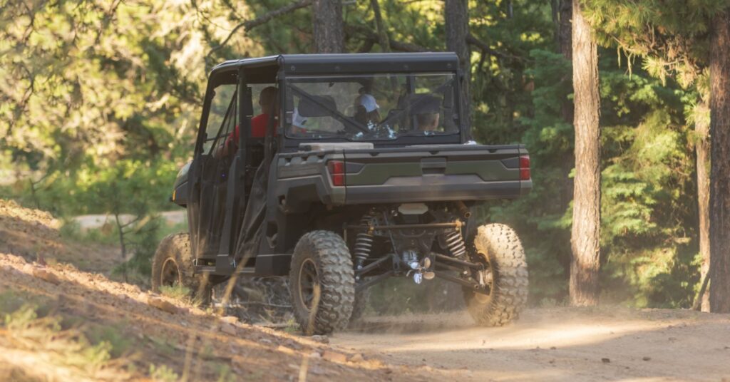 A dark green UTV carrying multiple passengers along a dusty dirt trail surrounded by tall trees in a forest.