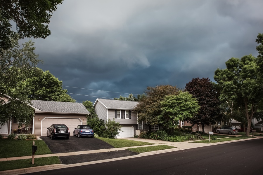 Dark thunderstorm clouds are spreading over a suburban neighborhood with houses, trees, and streets below.