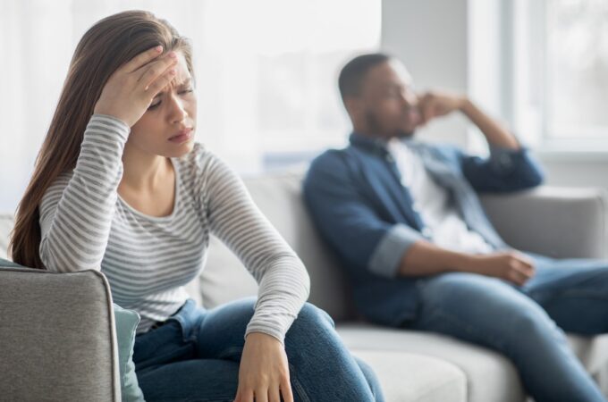 Woman looking distressed while sitting apart from her partner on a couch, suggesting tension or conflict in their interracial relationship.