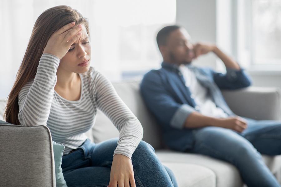 Woman looking distressed while sitting apart from her partner on a couch, suggesting tension or conflict in their interracial relationship.