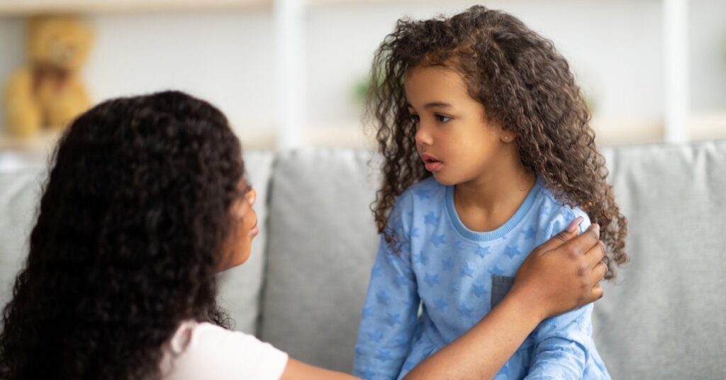 A young girl sits on a sofa and looks at a woman who kneels in front of her. The woman's hand is on the girl's shoulder.