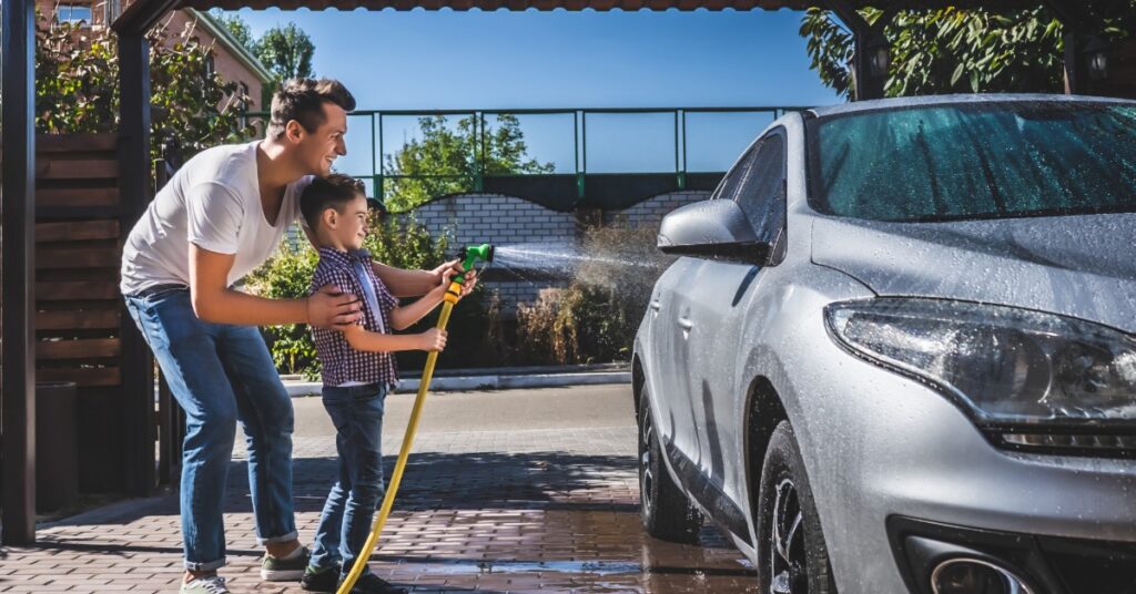 A man and his son are using a spray hose to clean the exterior of a car. They are in a driveway.