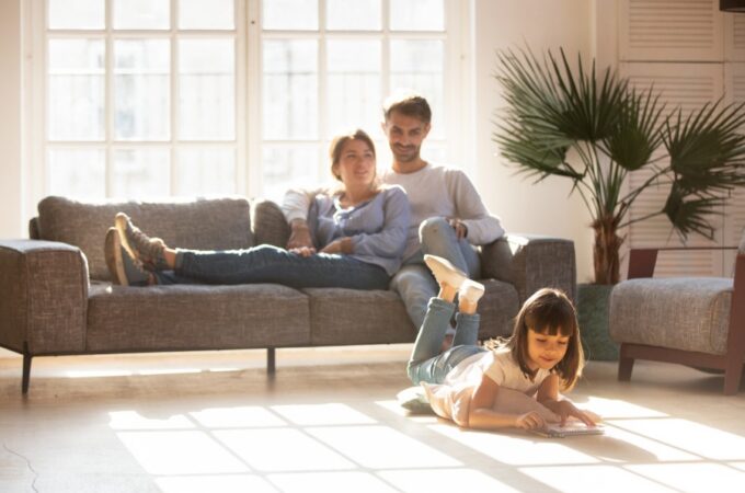 A young girl lying on the floor drawing in a notebook while two adults relax together on a gray sofa behind her.