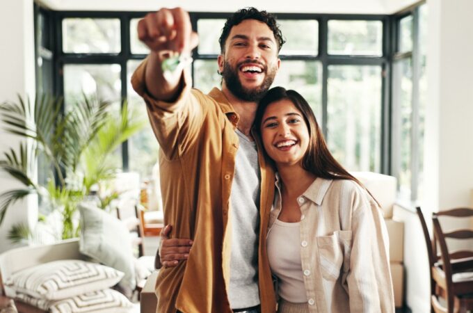 Two smiling people standing in a bright room, with one person holding keys forward and boxes behind them.