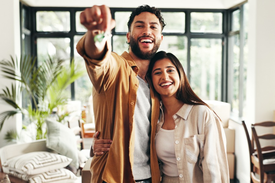 Two smiling people standing in a bright room, with one person holding keys forward and boxes behind them.