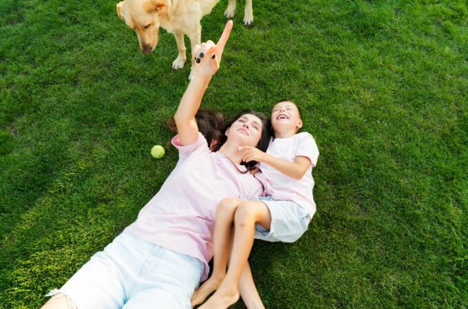 A close-up of a woman and a boy lying on grass, with a golden retriever standing nearby and a tennis ball beside them.