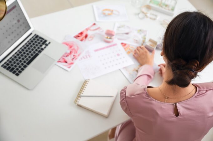 Woman in a pink blouse reviewing wedding planning materials and a calendar at a white desk with a laptop and notebook.
