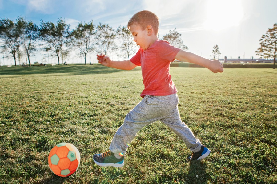 A young boy in a red shirt and gray pants kicks a bright orange and green soccer ball across a grassy field.