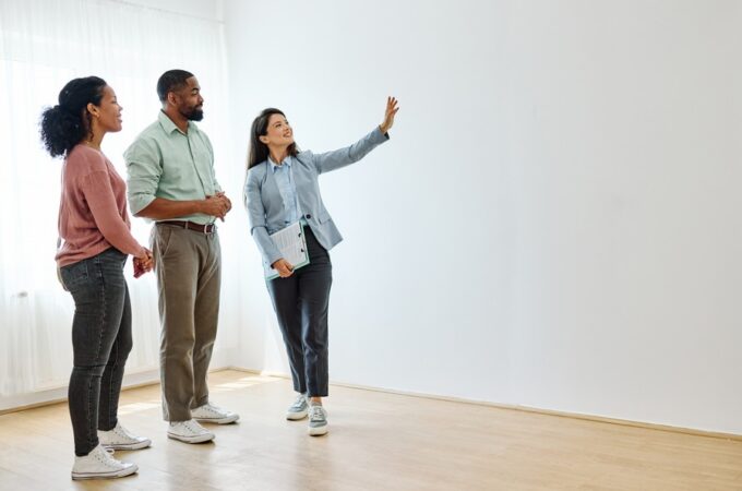 A woman holding a clipboard and gesturing toward a blank wall. Two people stand beside her in an empty room.