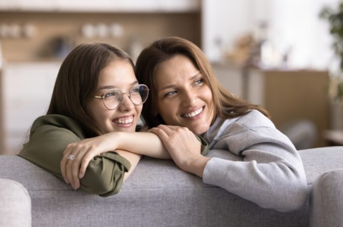 A woman in glasses and a green shirt shares a warm embrace with a smiling woman in gray on a living room sofa.