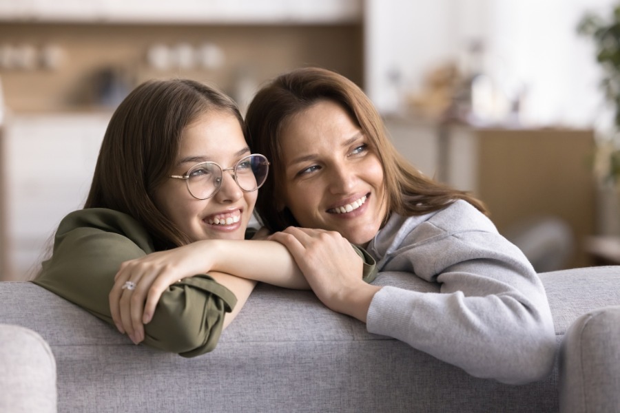 A woman in glasses and a green shirt shares a warm embrace with a smiling woman in gray on a living room sofa.