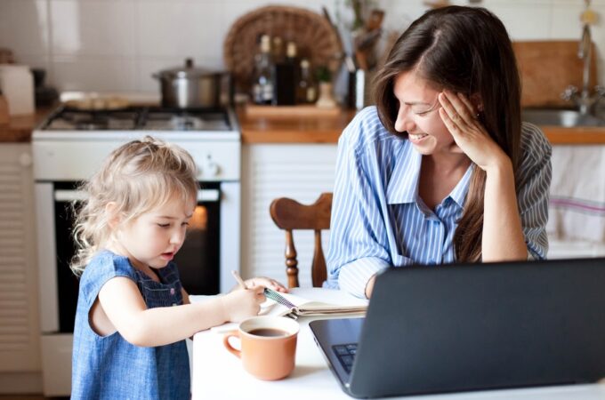 A smiling mother watches her young daughter write in a notebook at the kitchen table, with an open laptop and coffee mug nearby.