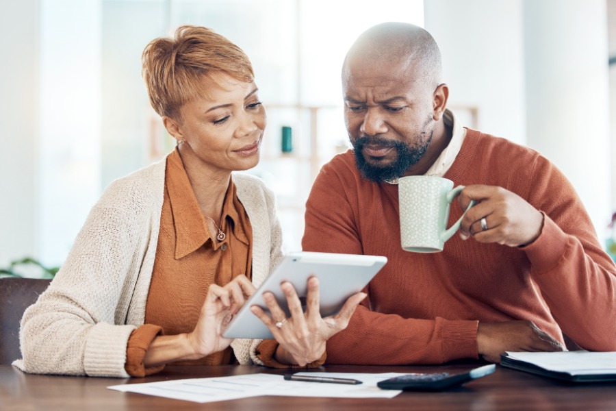 A man and a woman sit at a table looking at a table that the woman is holding. The man is holding a coffee mug.